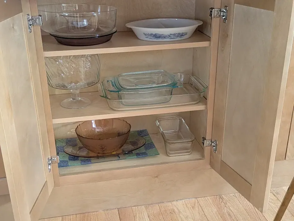 Open natural wood base cabinet with two shelves holding glass bakeware.