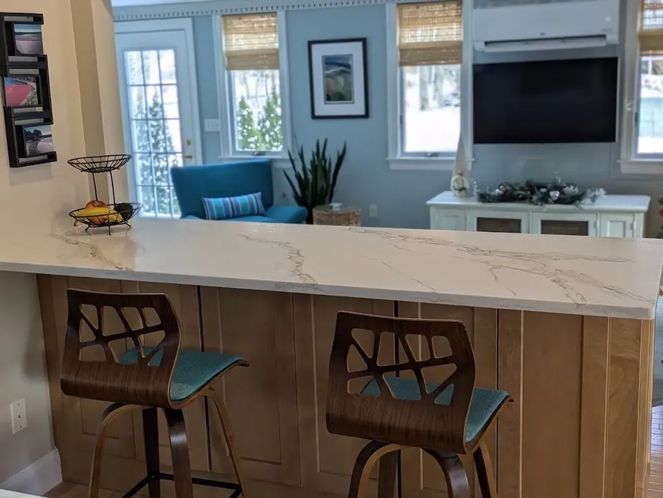 Peninsula with white veined countertop and two stools, sitting room with windows beyond.