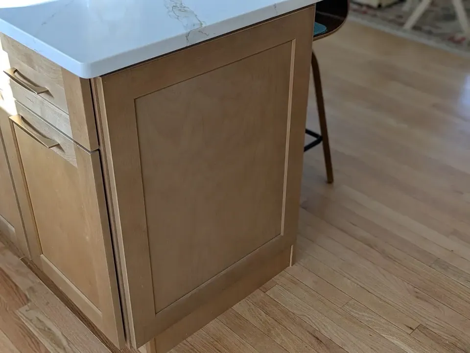 Natural wood shaker end panel and drawers under the white countertop edge, wood floor.