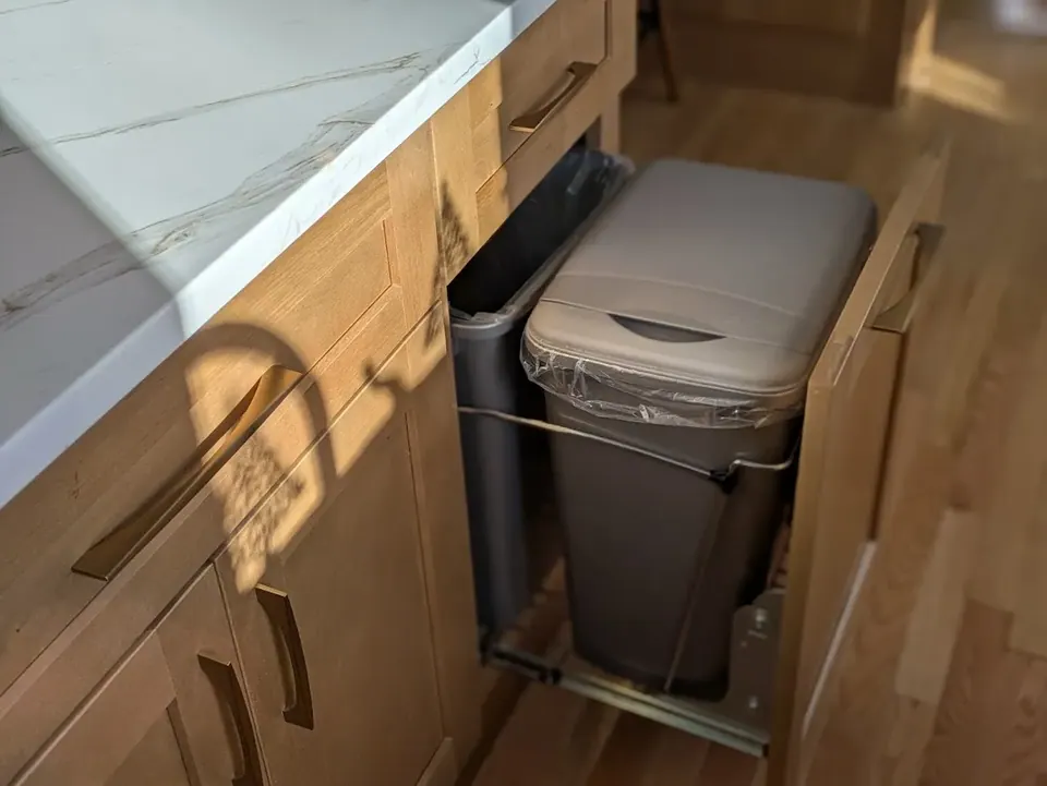 Pull-out trash bin open in a natural wood base cabinet under the white countertop.