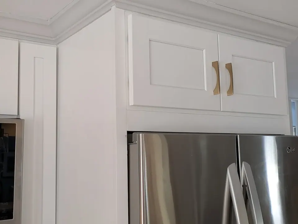 White shaker cabinet above the refrigerator with gold pulls and crown molding.
