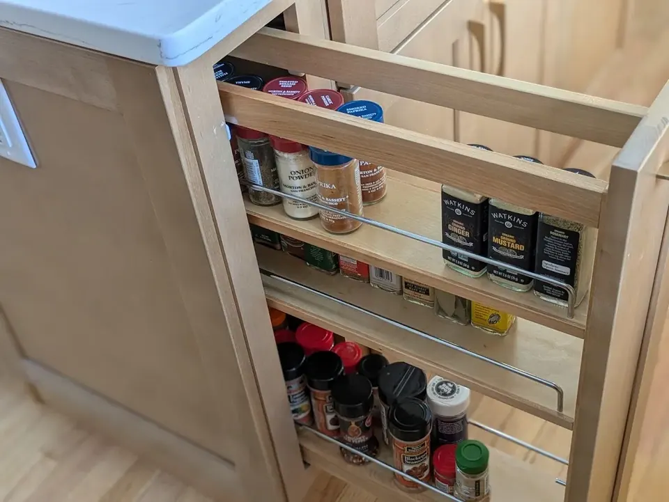 Pull-out spice rack with three shelves of jars in a natural wood base cabinet.