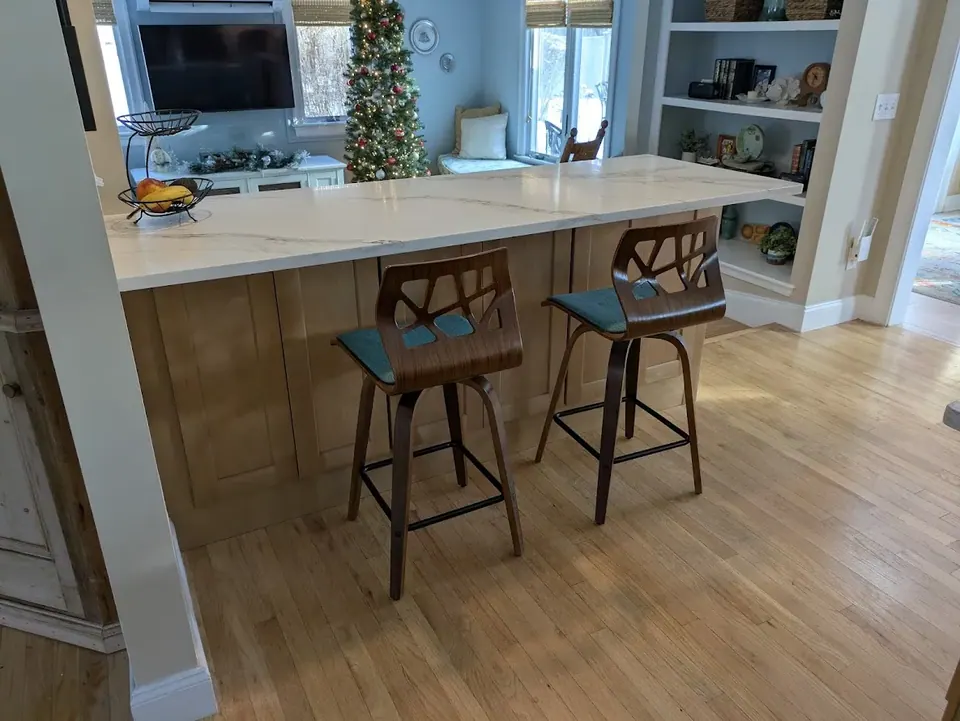 Peninsula with white veined countertop and natural wood panels, two stools, living room beyond.