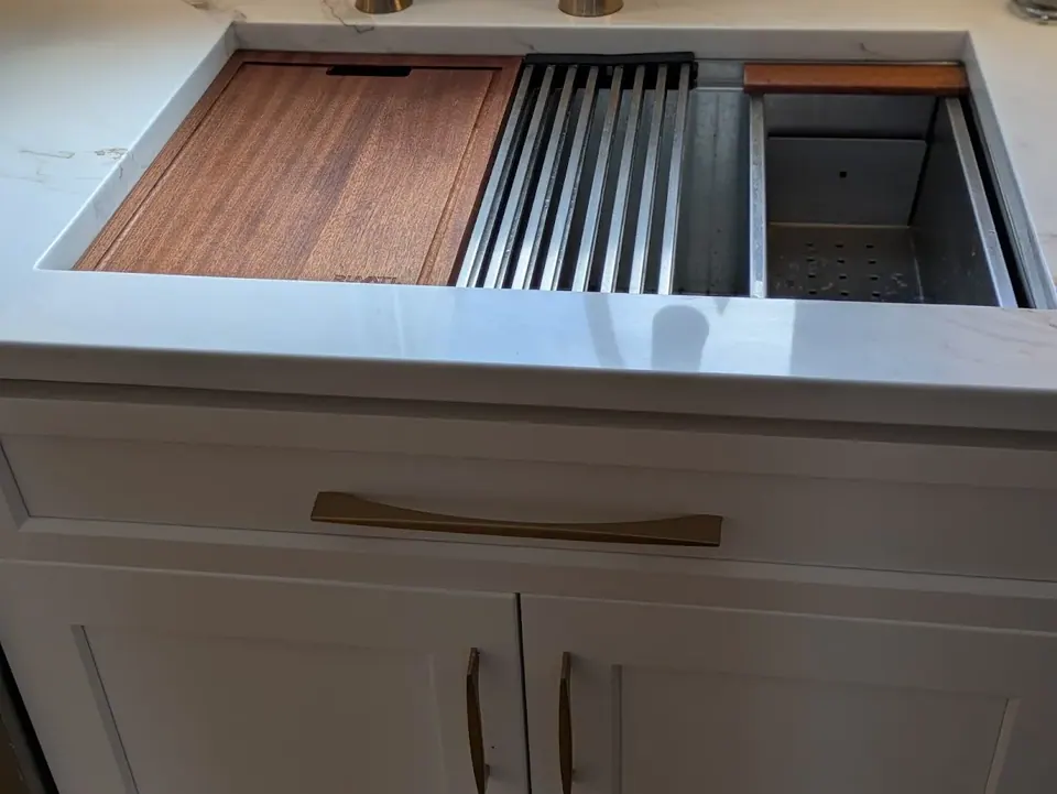 Sink with cutting board and drying rack above a white sink base with gold pulls.