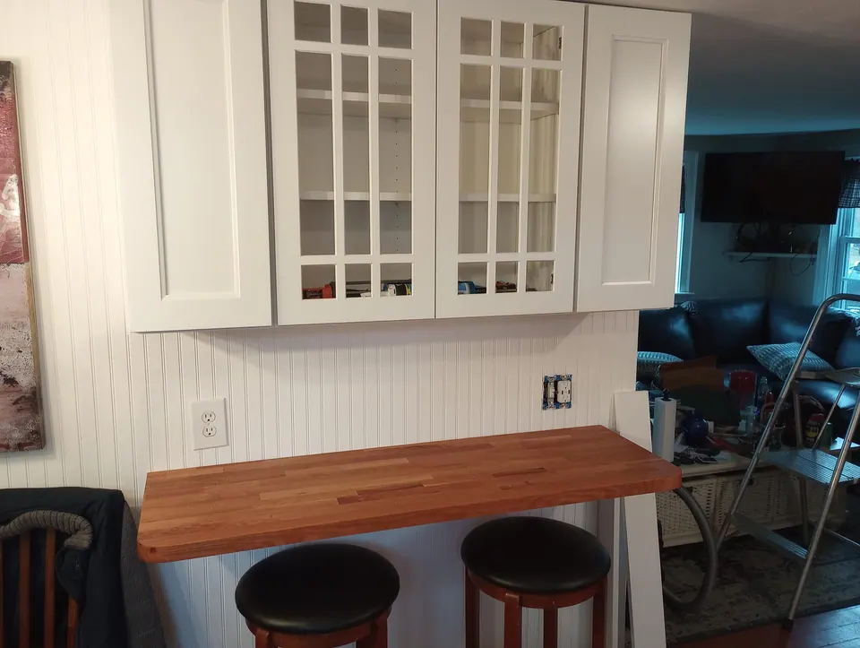 White wall cabinets with two glass mullion doors above a butcher-block bar top and two stools.