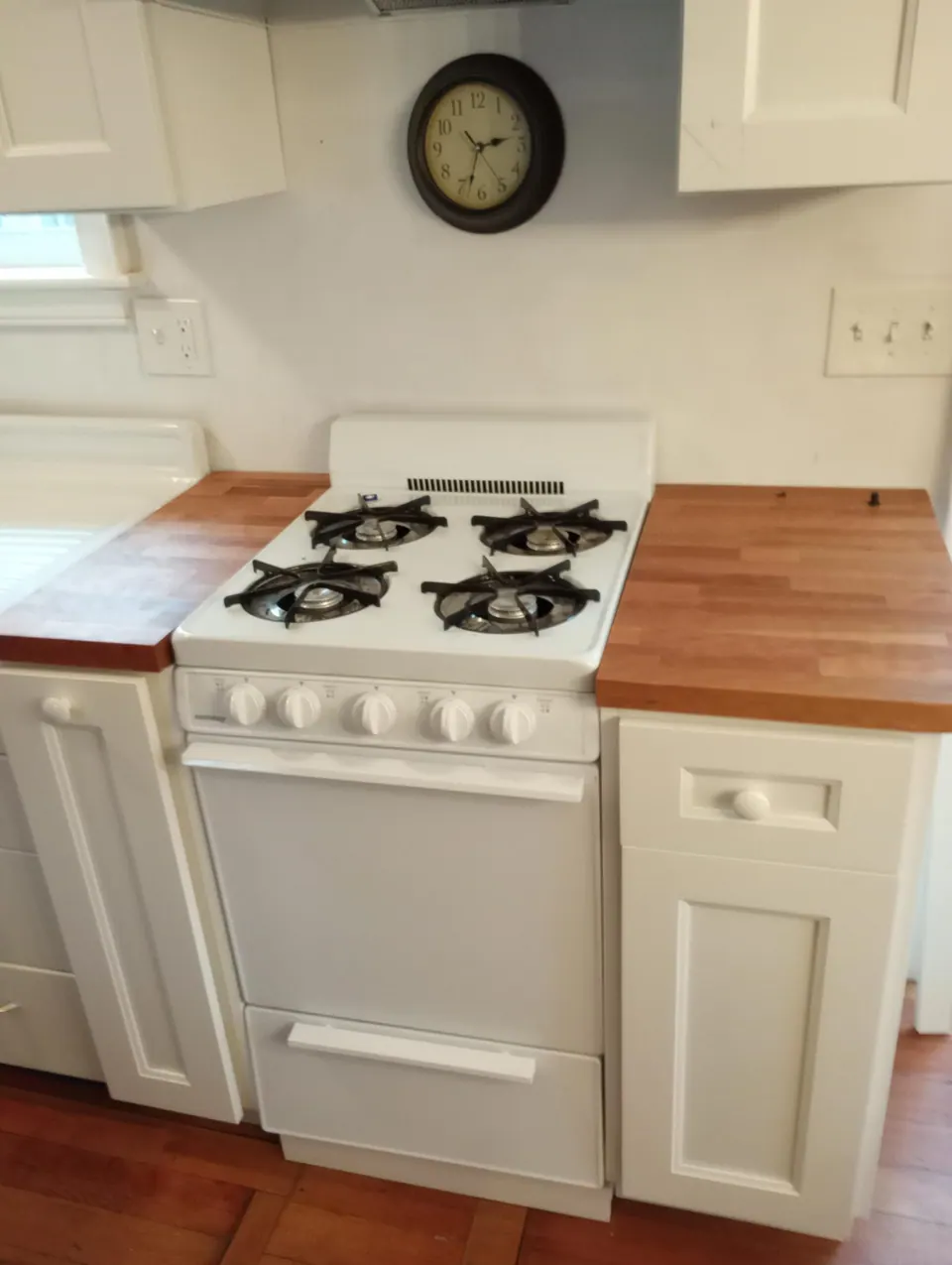 White gas range between white base cabinets with round knobs and butcher-block counters, wall clock above.