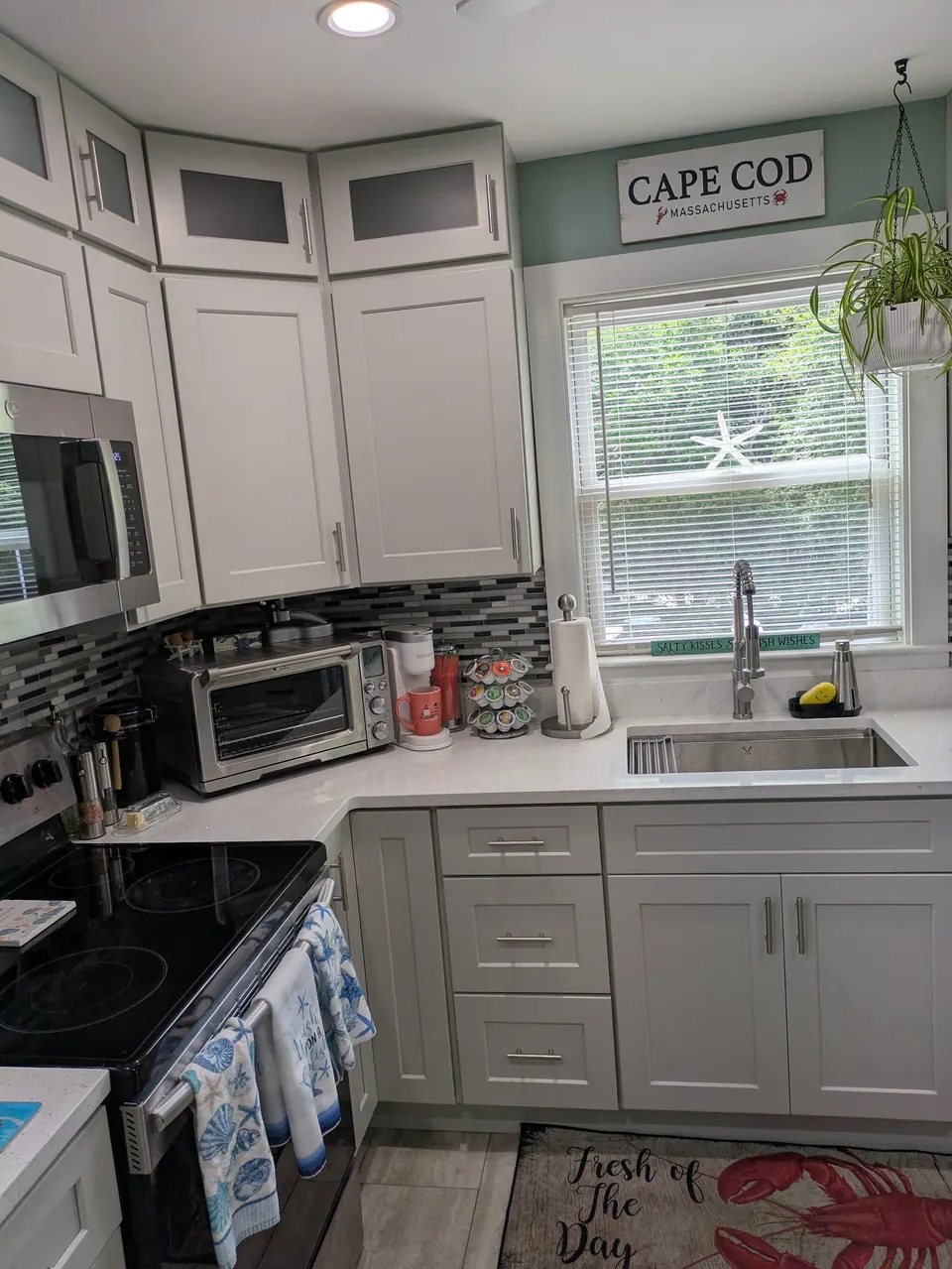 Corner of white shaker cabinets with a sink under the window and a Cape Cod sign.