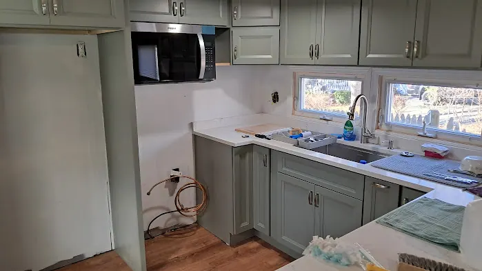 Sage green cabinets with white countertop, sink under two windows, microwave above the empty range space.