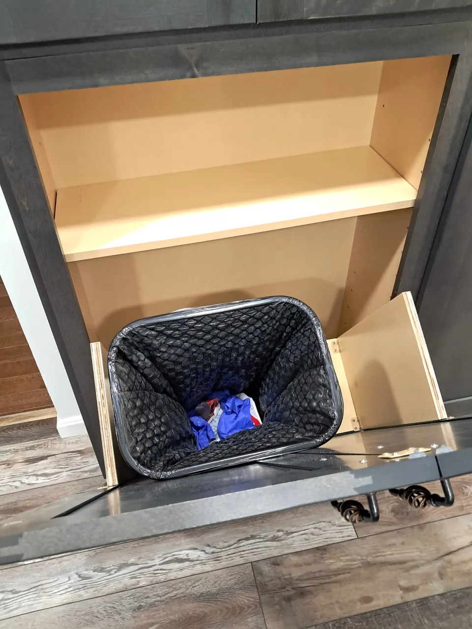 Tilt-out trash bin open at the base of the dark gray pantry cabinet, shelf above.