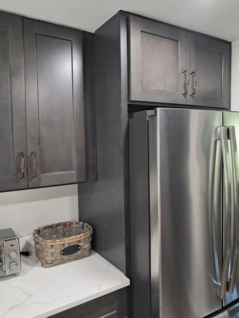 Dark gray wall cabinet and over-refrigerator cabinet beside a stainless refrigerator and white veined countertop.