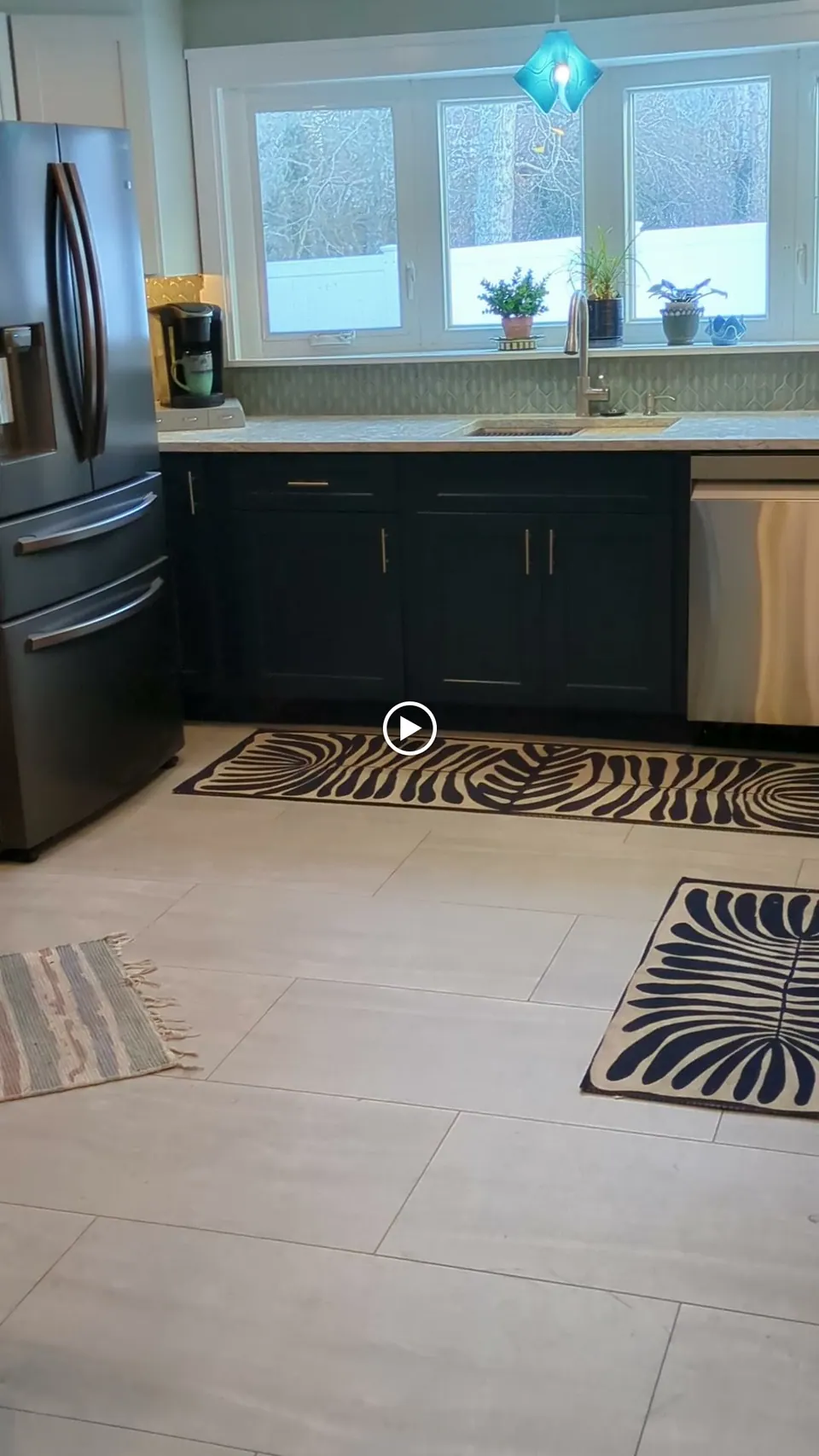 Kitchen view with navy blue base cabinets, sink under the window, dark refrigerator and light tile floor.