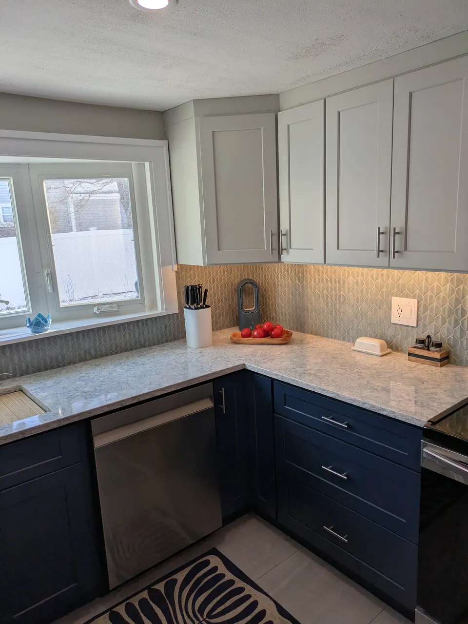 Light gray wall cabinets over navy blue base cabinets and a dishwasher in the corner by the window.
