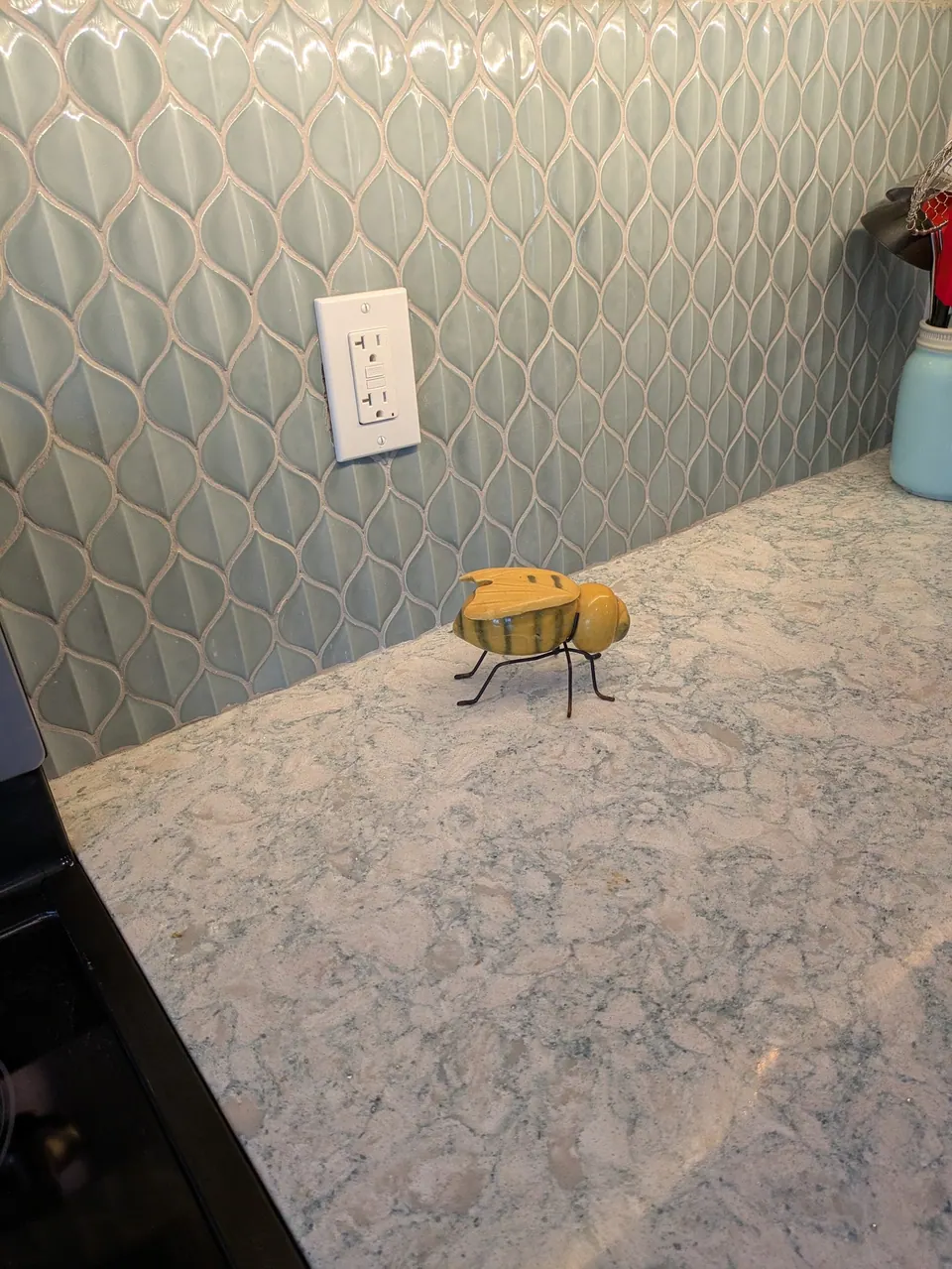 Glossy pale green leaf-pattern backsplash tile behind the cream veined countertop, with a small bee ornament.