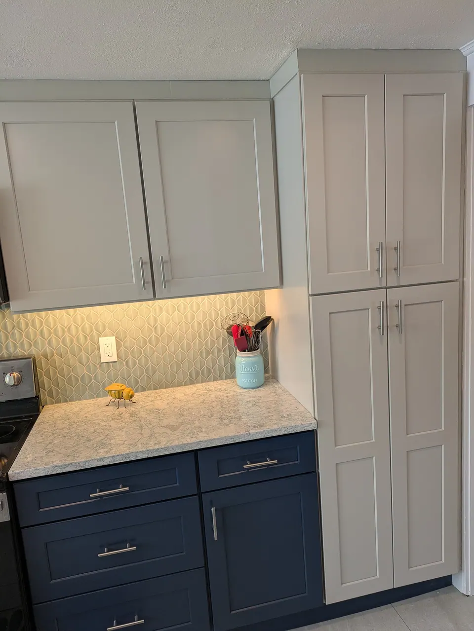 Light gray wall cabinets and tall pantry cabinet beside navy blue drawer bases with under-cabinet lighting.