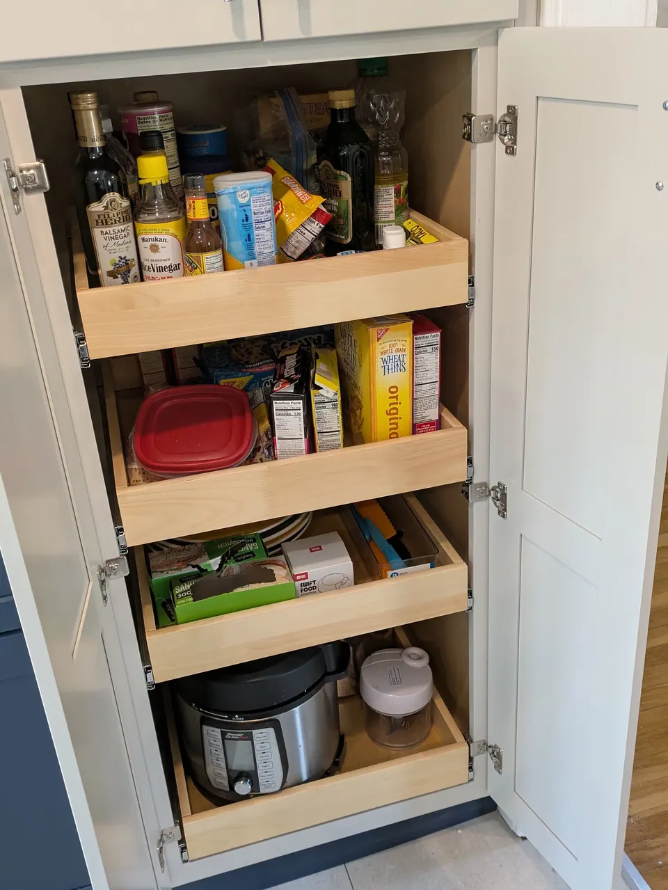 Light gray pantry cabinet open, showing four roll-out trays stocked with food and a pressure cooker.