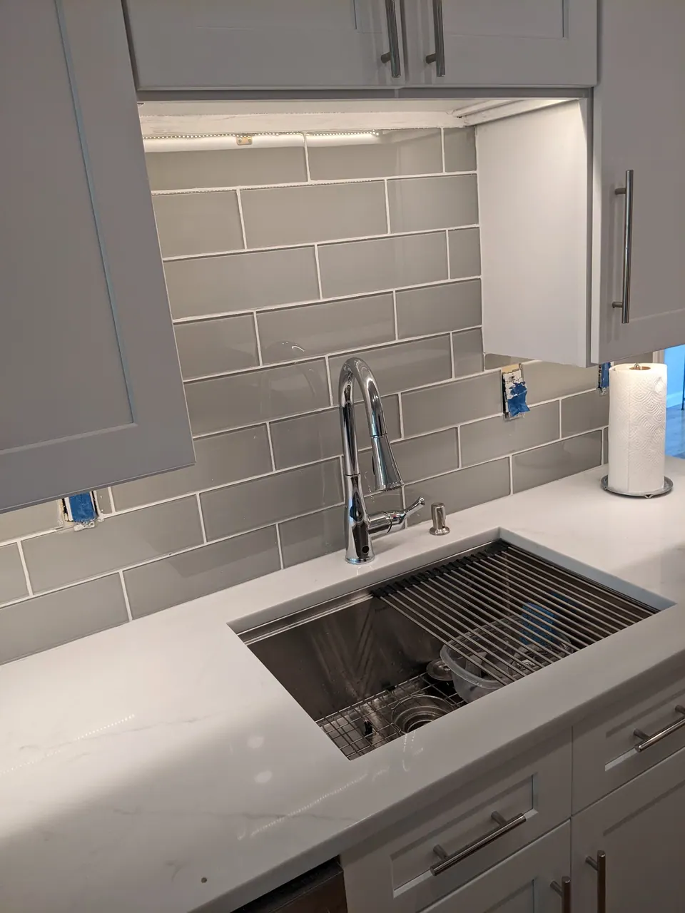 Workstation sink under lit wall cabinets and a gray glass tile backsplash.