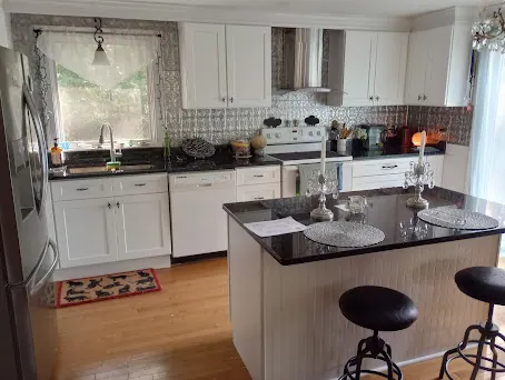 White shaker kitchen with island, two black stools, black speckled countertop and sink under a window.