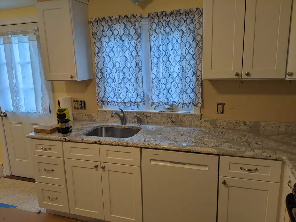 Sink run of white shaker cabinets under a curtained window, granite counter.