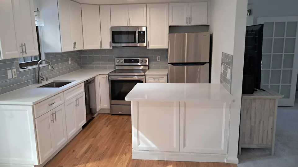 L-shaped white shaker kitchen with a quartz island, stainless appliances and a gray-blue tile backsplash.