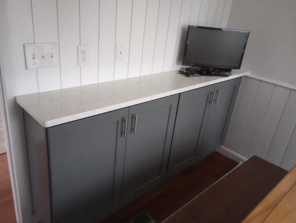 Gray shaker base cabinets with a white countertop along a white paneled wall, small TV on top.