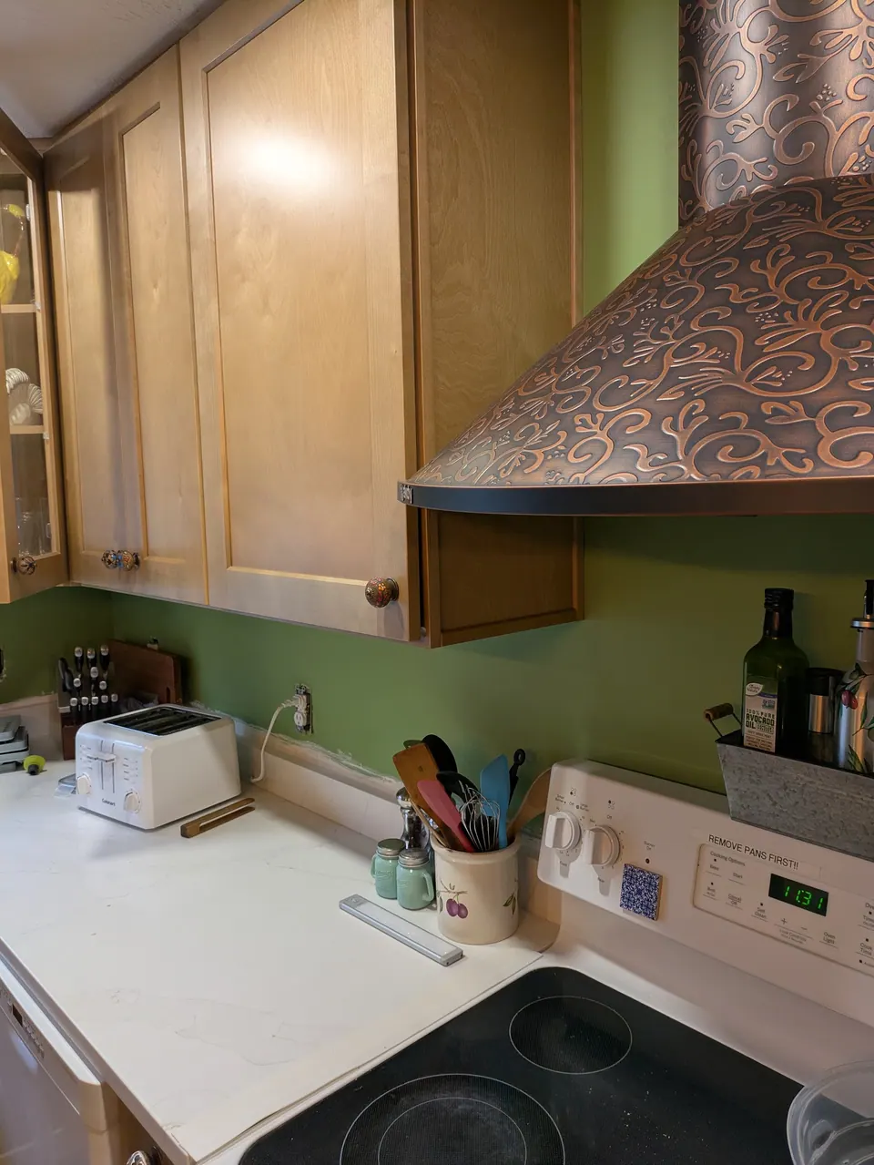 Light wood wall cabinets with patterned knobs beside the copper-tone hood, white countertop and cooktop below.