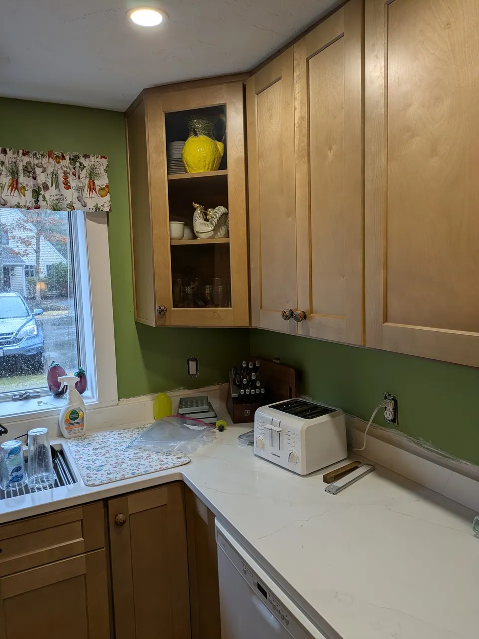 Glass-front corner wall cabinet and light wood wall cabinets above the white countertop near the window.