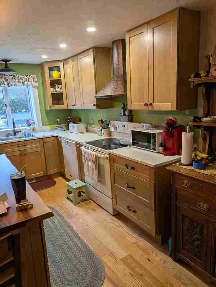 Light natural wood shaker cabinets with a copper-tone range hood, white range and white countertop, wood floor.