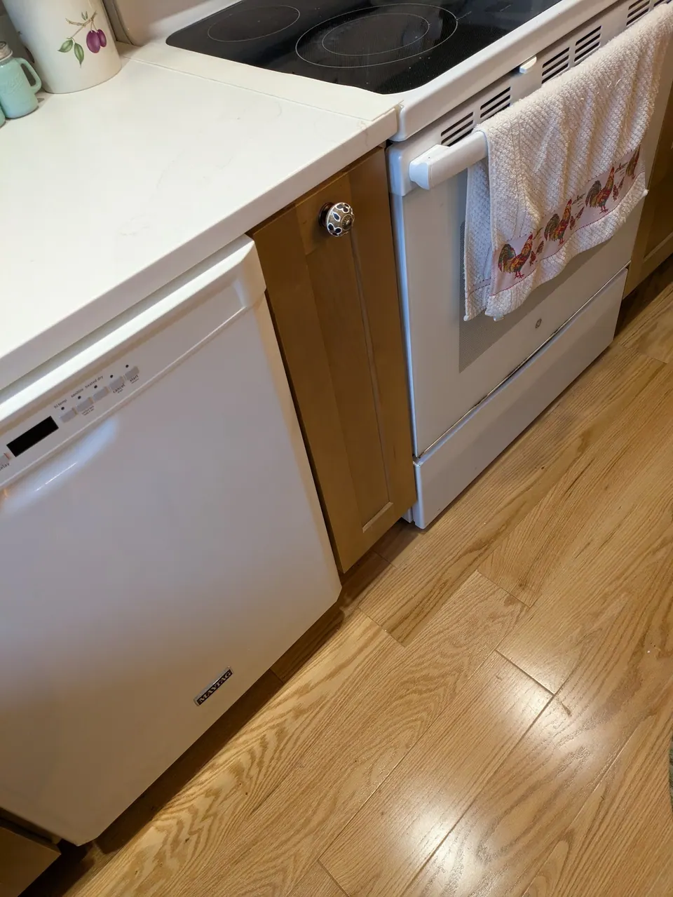 Narrow light wood cabinet front with a round knob between the white dishwasher and range.