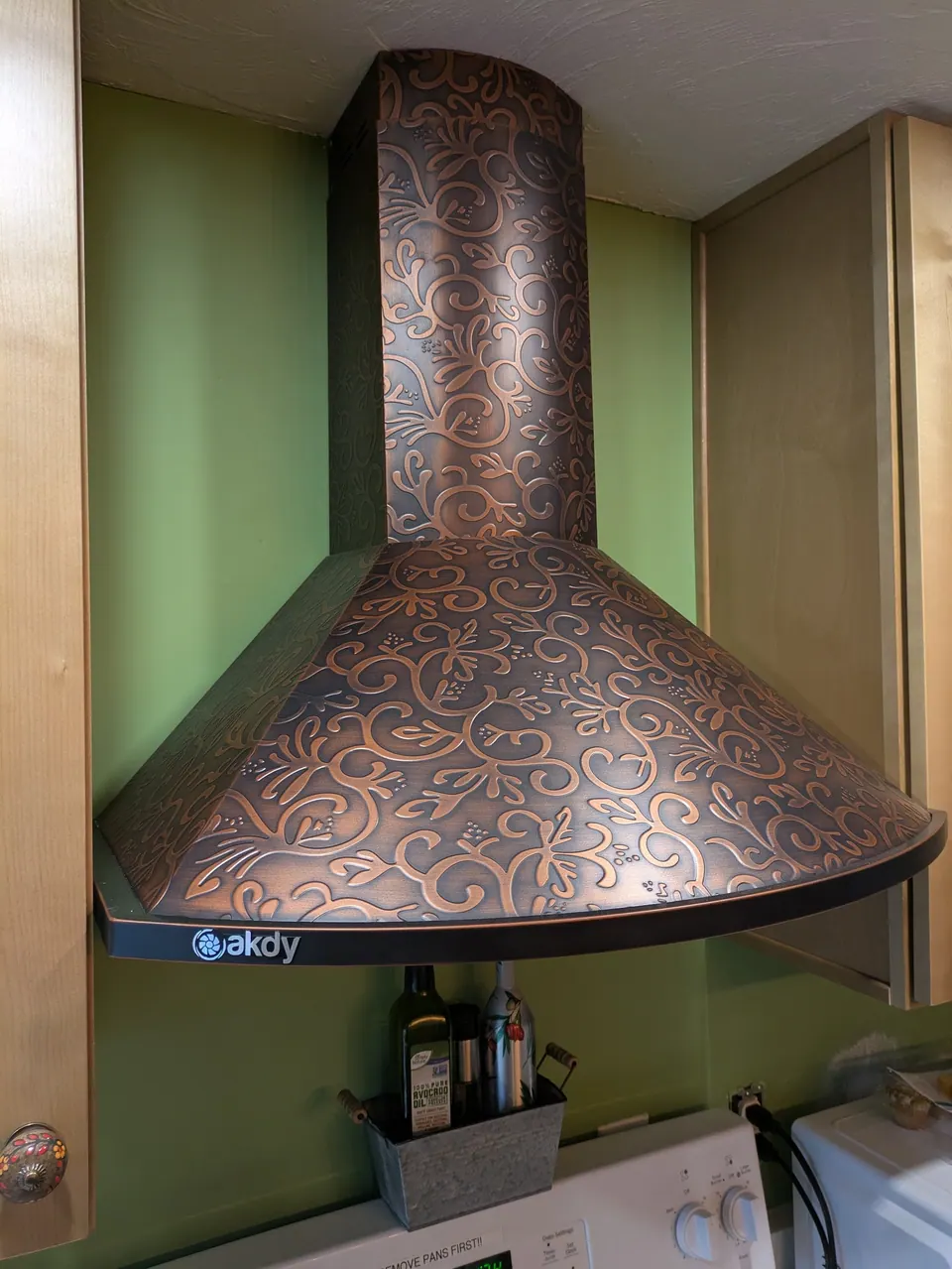 Embossed copper-tone chimney range hood against a green wall between light wood cabinets.