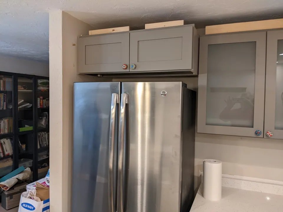 Refrigerator between tall gray cabinets with glass-front cabinets above.