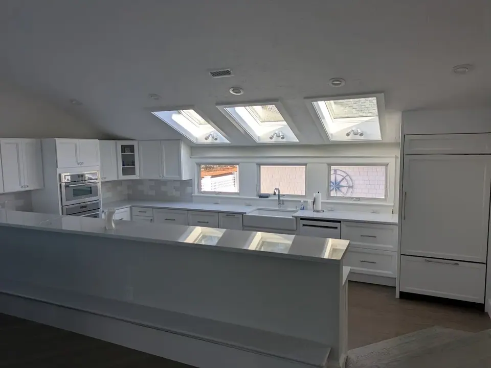 Whole white kitchen under three skylights, with wall ovens, farmhouse sink and paneled refrigerator.
