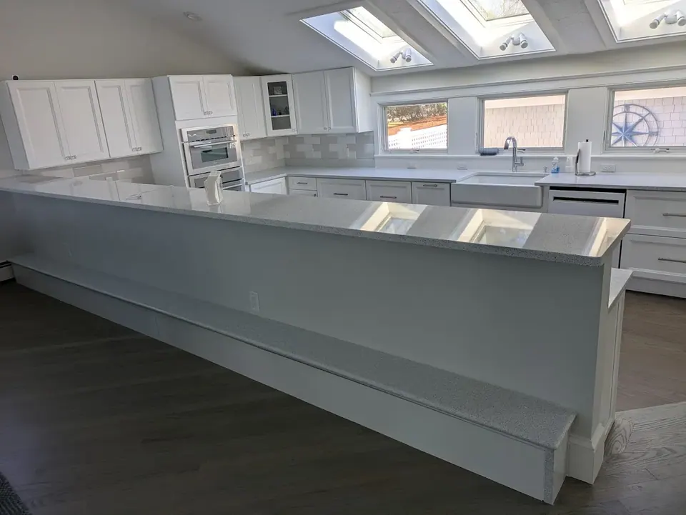 White kitchen behind a long half wall with speckled countertop, wall ovens, farmhouse sink and skylights.