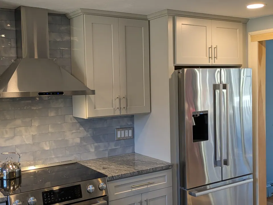 Stainless chimney hood and refrigerator framed by light gray cabinets with crown molding.
