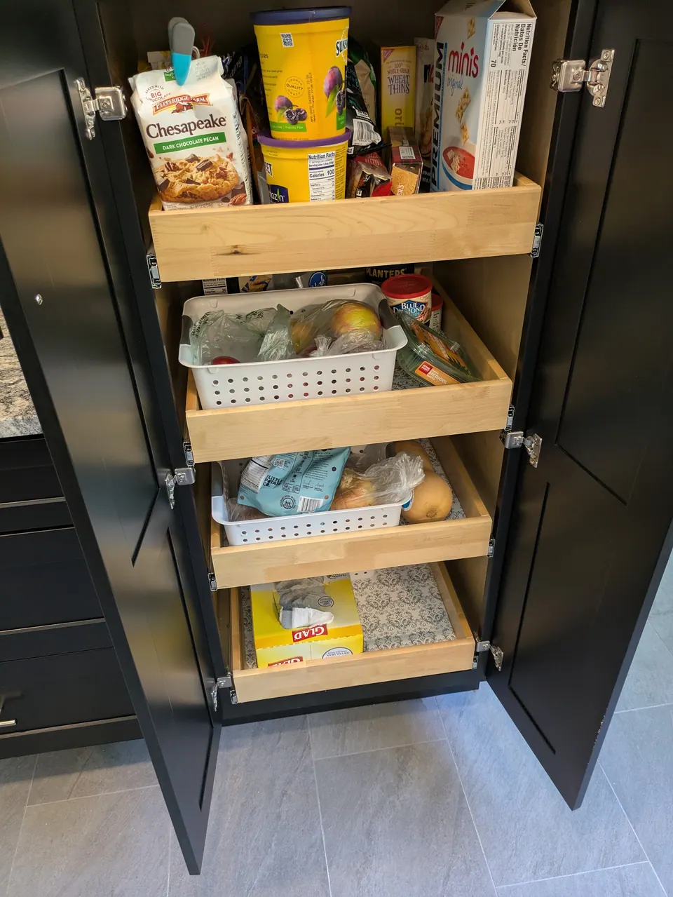 Tall black pantry cabinet open, showing four wood roll-out trays stocked with food.