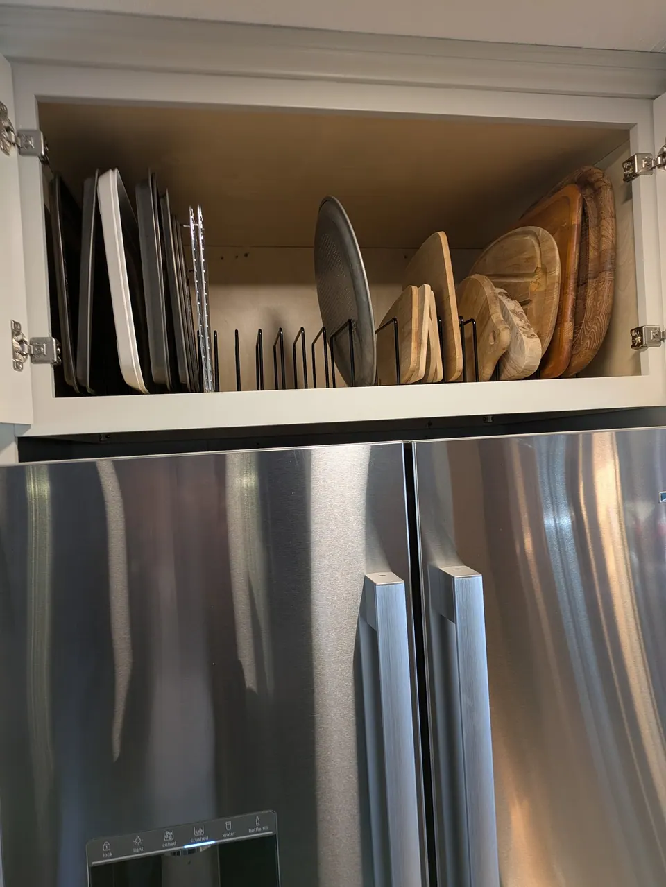 Open cabinet above the refrigerator with a wire divider holding baking sheets and boards.
