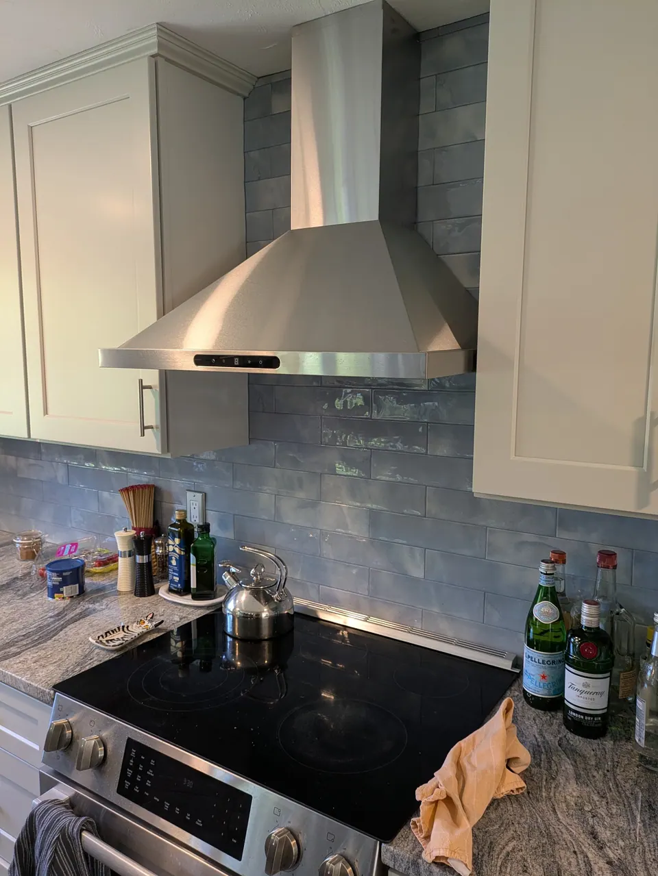Stainless chimney hood between light gray wall cabinets over a glossy blue tile backsplash.