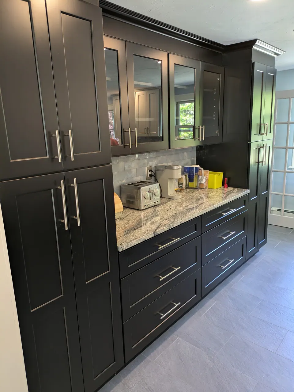 Black shaker pantry wall with glass-front upper cabinets, drawer bases and gray veined countertop.
