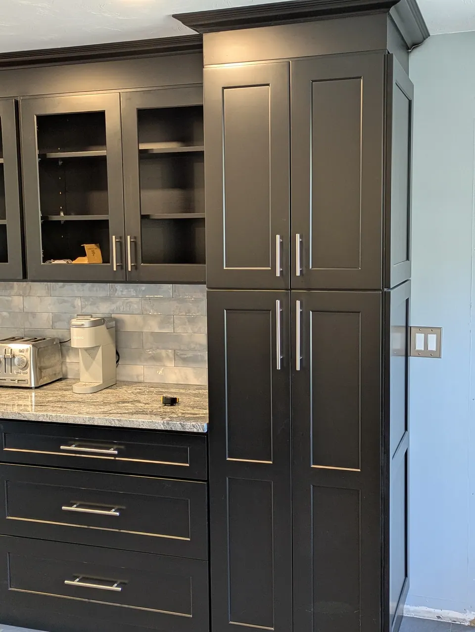 Tall black pantry cabinet beside empty glass-front wall cabinets and a black drawer base.