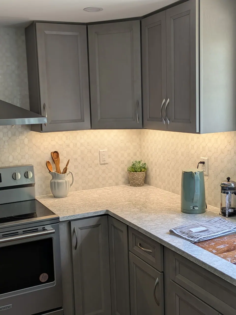 Gray wall and base cabinets around a corner, with range, light countertop and lit mosaic backsplash.