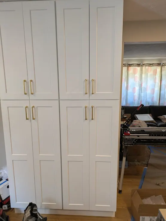 Wall of tall white shaker pantry cabinets with brass-tone bar pulls on a wood floor.