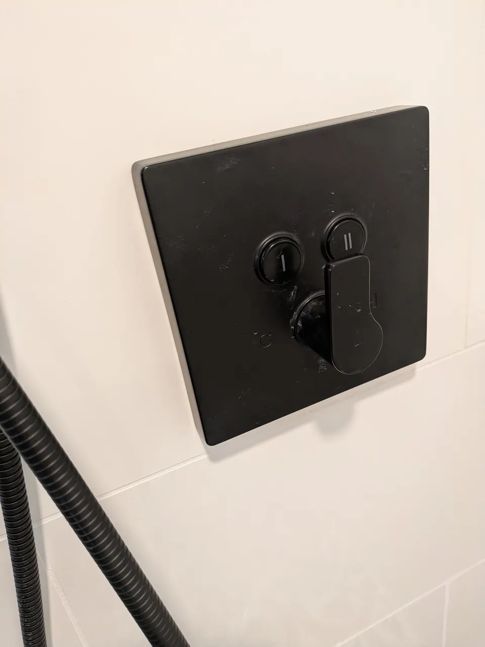 Square matte black shower valve trim on a white tiled wall.