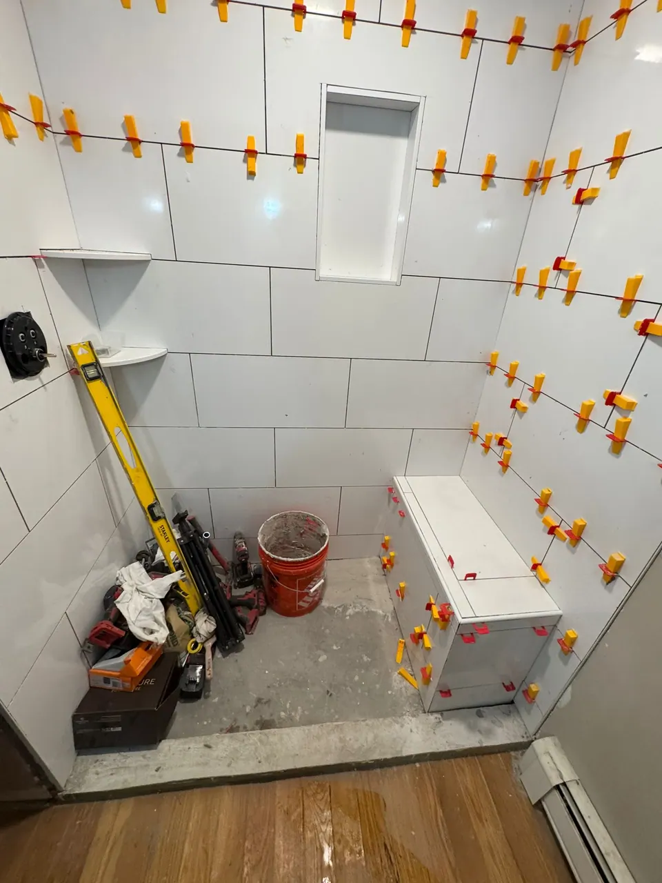 Shower being tiled: white wall tile held by leveling clips, bench, niche and tools on the floor.