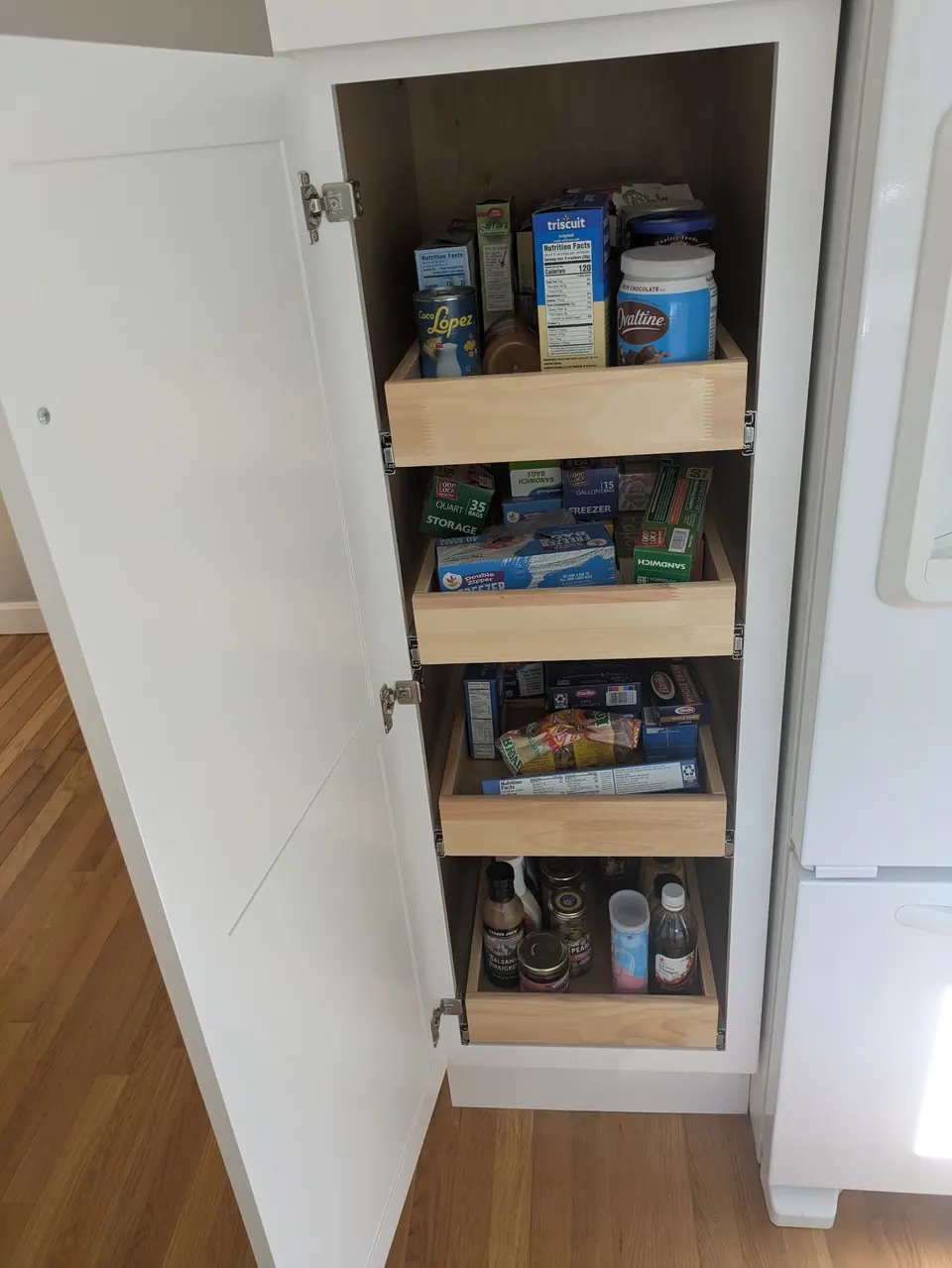 White pantry cabinet open beside the refrigerator, showing four wood roll-out trays stocked with food.