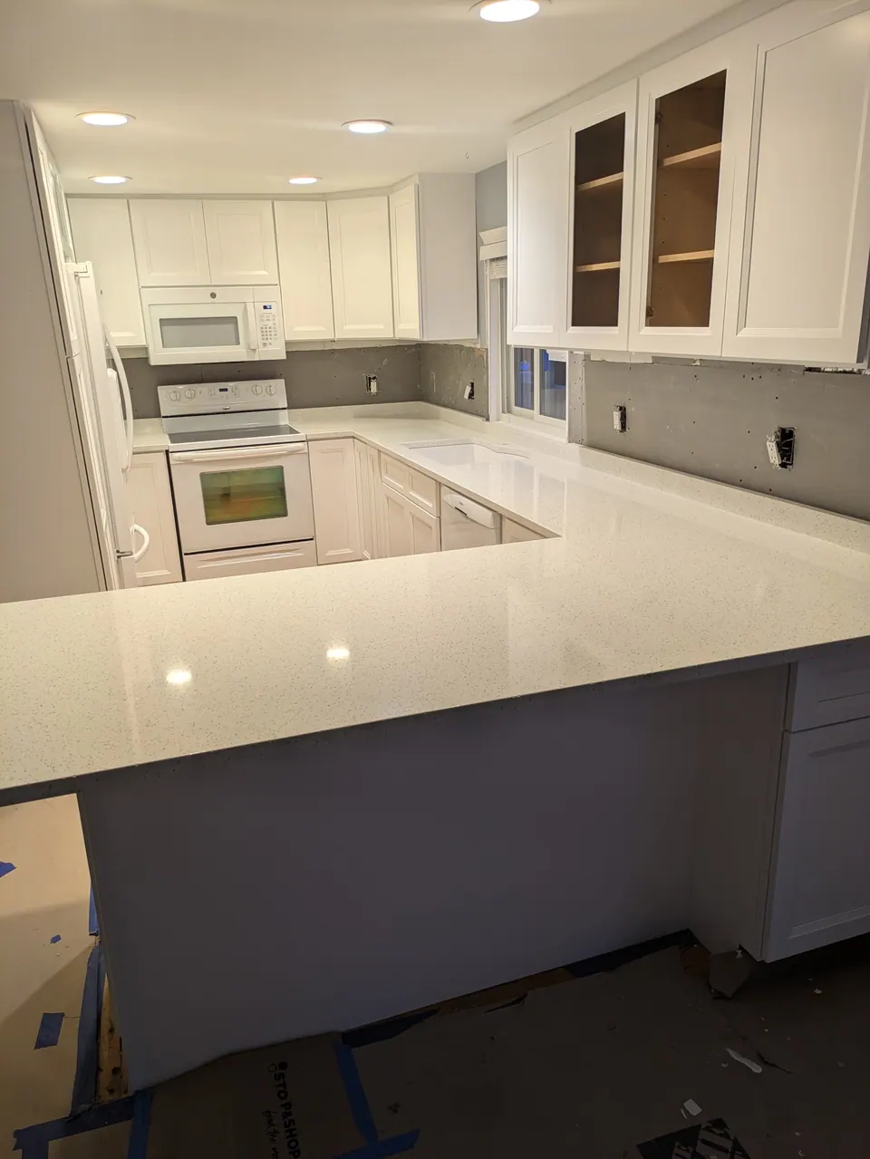 In progress: U-shaped white kitchen with peninsula countertop installed, bare wall before backsplash, floor covered.