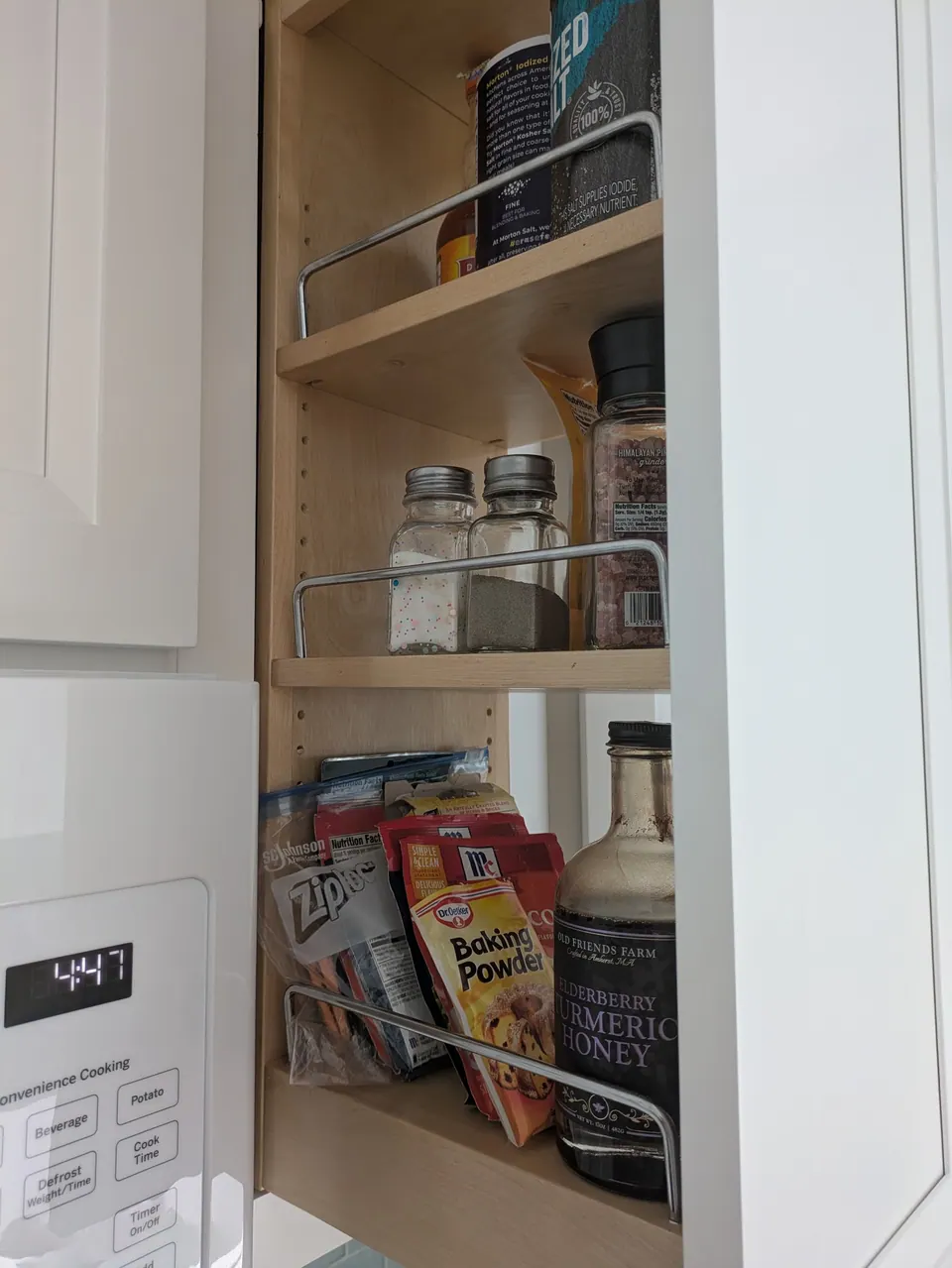 Narrow wall cabinet open beside the microwave with chrome-railed shelves holding spices and baking supplies.