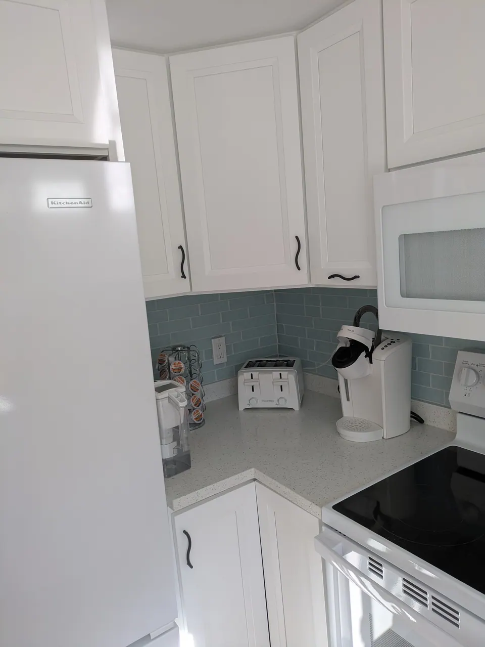 White corner wall and base cabinets with black pulls between the refrigerator and range, light blue backsplash.