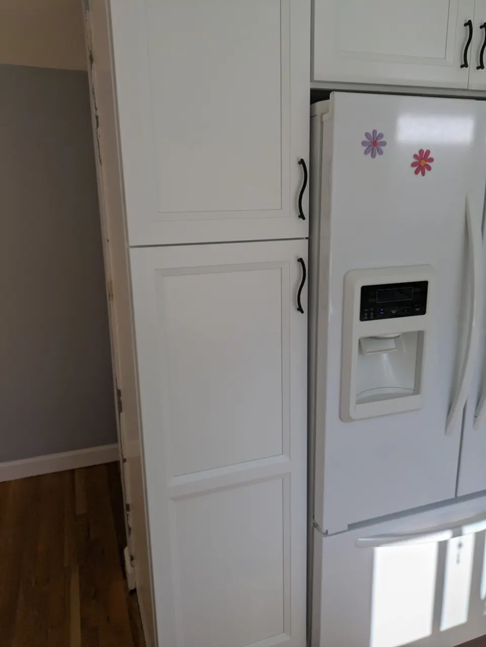 Tall white pantry cabinet with two doors and black pulls beside the white refrigerator.