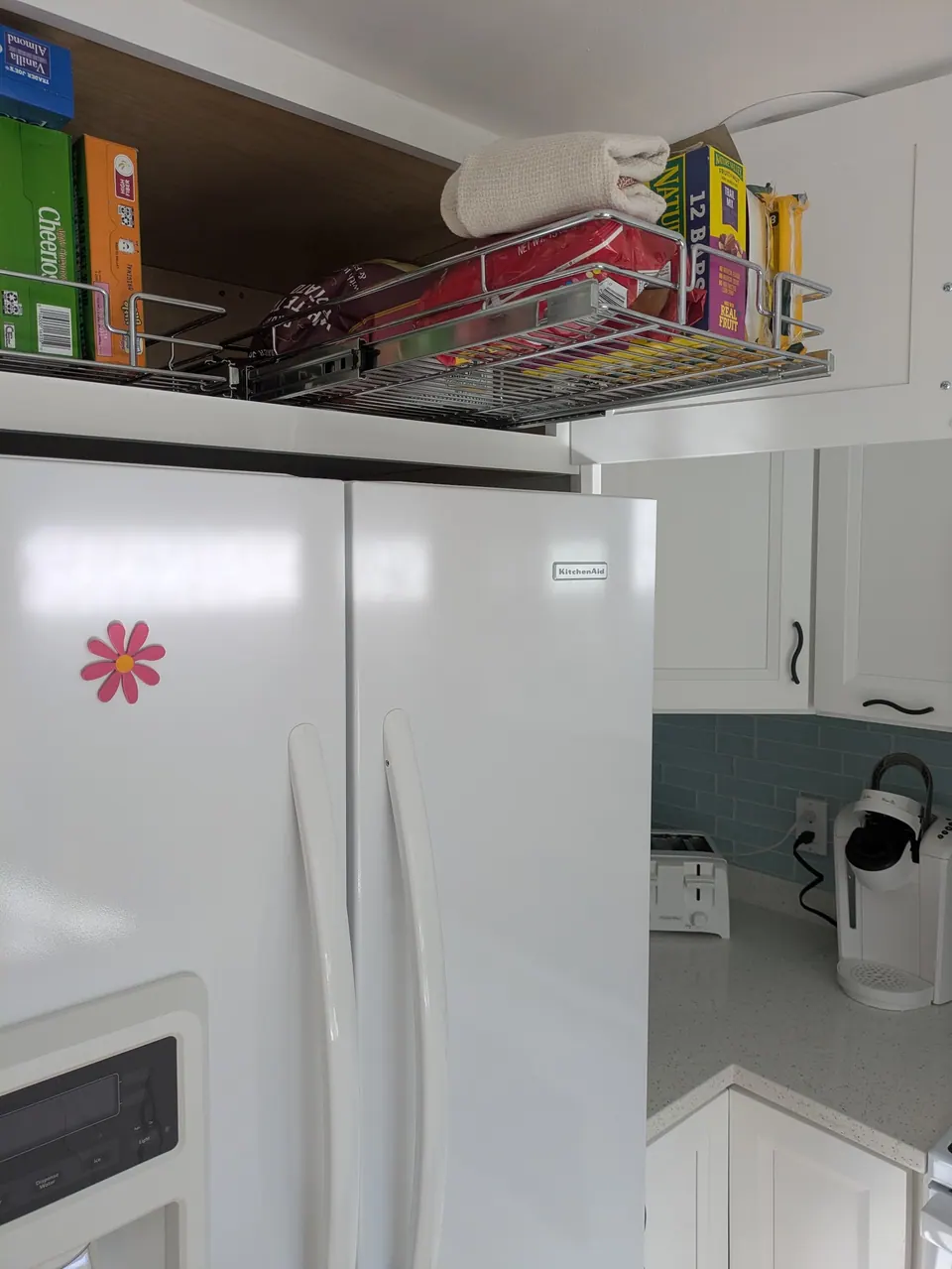 Wire pull-out basket extended from the cabinet above the white refrigerator, holding boxed food.