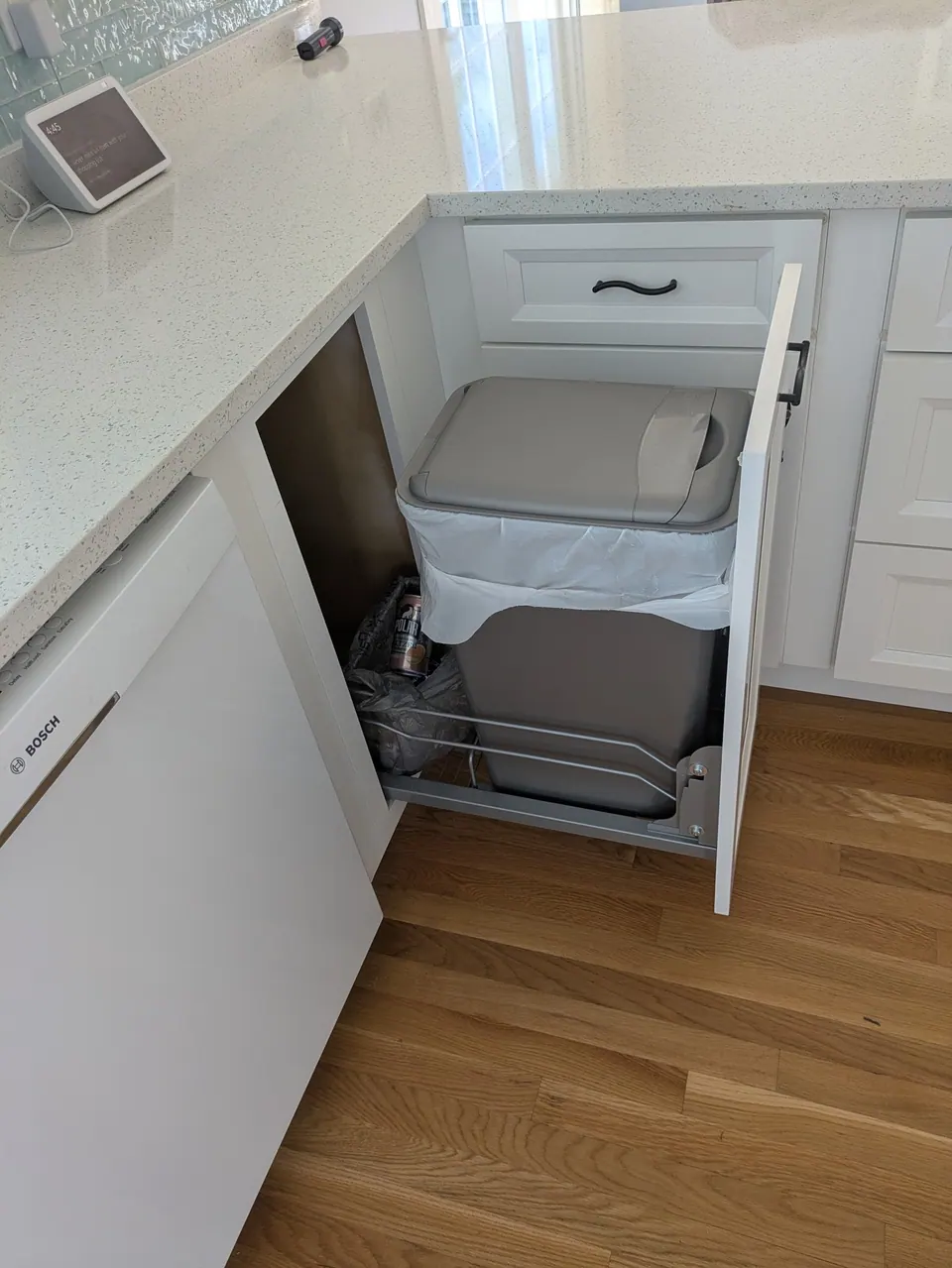 Pull-out trash bin open beside the dishwasher under the speckled white countertop.