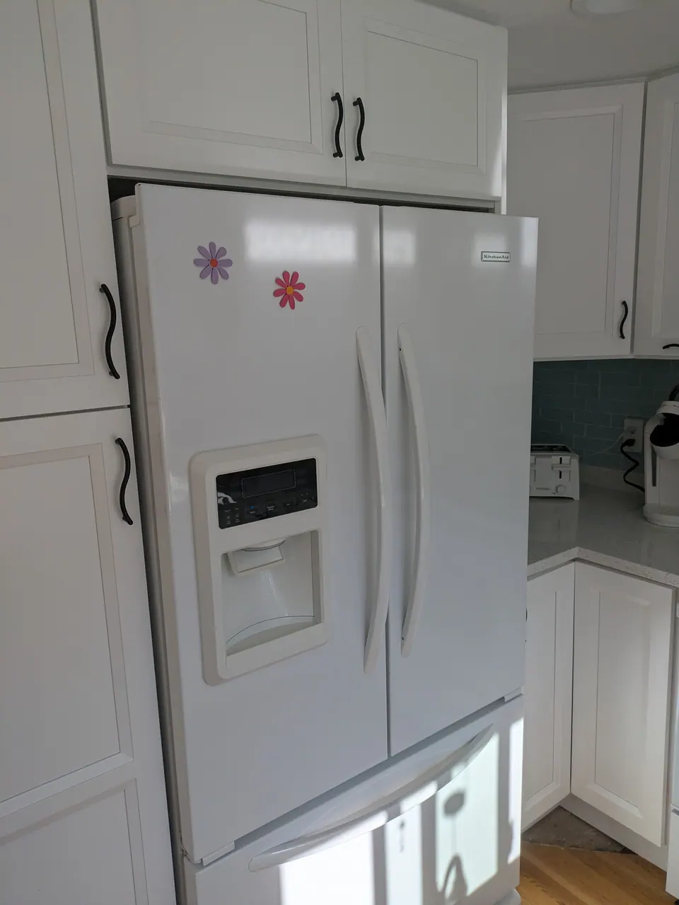 White refrigerator framed by white shaker cabinets, with a cabinet above and tall pantry doors beside it.